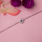 A silver-colored bracelet with a blue evil eye charm, displayed on a pink background with a purple flower and pink petal in the corner.